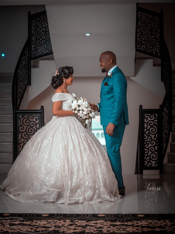 Groom In A Teal Suit Holding Hands With A Bride In A Massive, Shimmering White Ballgown In A Modern Hall.